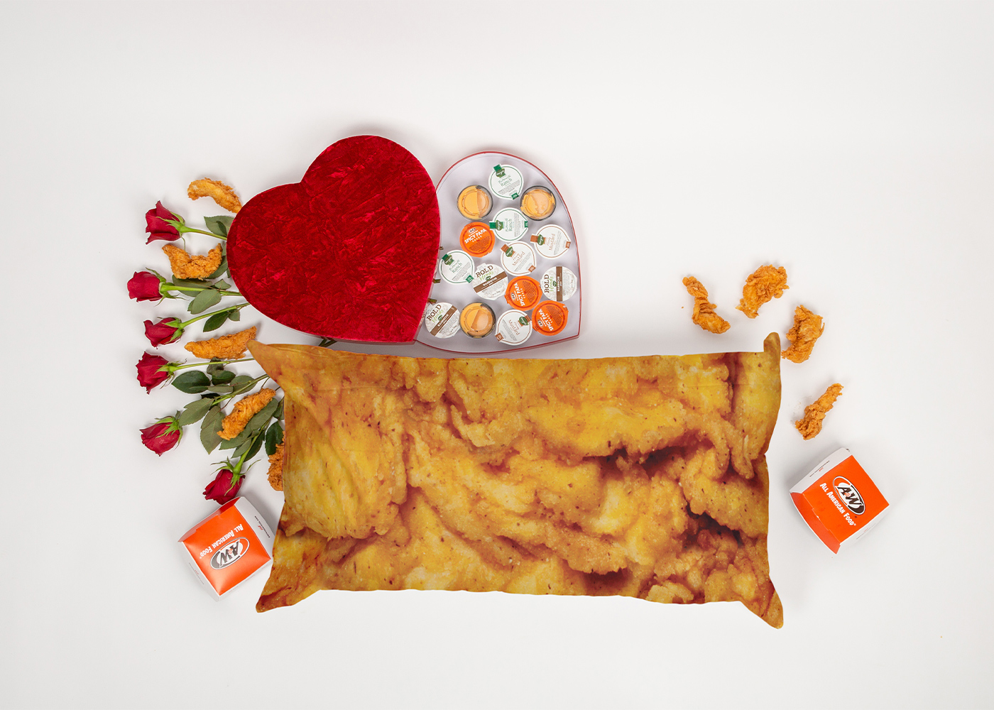 Flat lay photo of a body pillow that looks like an A&W Chicken Tender. The pillow is surrounded by a heart-shaped box filled with A&W sauces and Chicken Tenders.